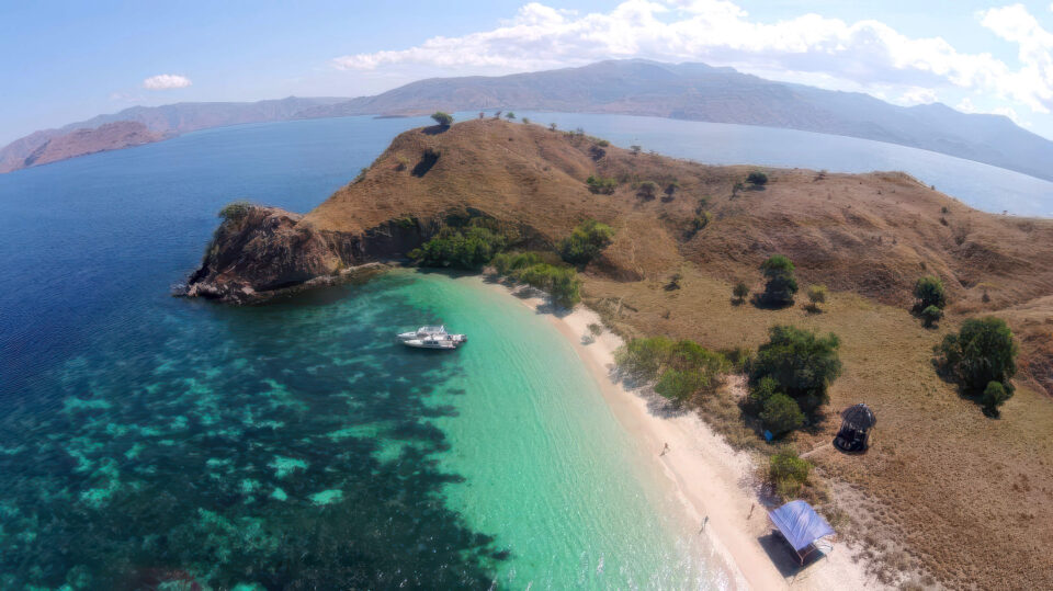 Vue drone sur une plage de sable blanc isolée, entourée de collines arides et d'eaux turquoise, avec un bateau ancré au large lors d'une croisière Komodo Indonésie